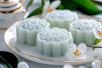 Delicate mint green mooncakes with floral pattern served on elegant porcelain plate, garnished with fresh white blossoms and green leaves for Asian dessert presentation.