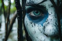 Close-up portrait of person with dramatic blue and white tribal face paint featuring intricate patterns around striking green eye in natural forest setting.