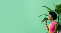 Young woman with dark curly hair and medium skin tone wearing bright pink top, standing against mint green wall with tropical green plant foliage, looking upward thoughtfully for lifestyle and wellness content.