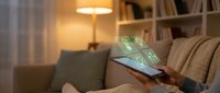 Person using smartphone with holographic financial interface displaying dollar symbols and digital banking icons in cozy living room setting.