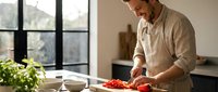 Young man in beige sweater chopping fresh red tomatoes in modern kitchen with natural light from large windows for healthy cooking and meal preparation.