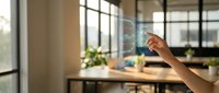 Young adult hand adjusting smart home temperature control on glass panel in modern office workspace with natural light and plants.
