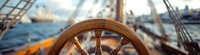 Wooden ship steering wheel on sailing vessel against blurred harbor background with boats and blue water, maritime navigation equipment.