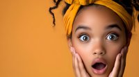 Young African American woman with natural curly hair wearing yellow headband expressing surprise with hands on cheeks against orange background.