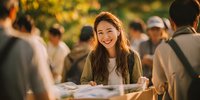 Young Asian woman smiling while holding tray at outdoor event with crowd in background during golden hour sunlight.