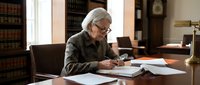 Senior Caucasian male lawyer with gray hair and glasses reviewing legal documents in traditional law office with leather-bound books and wooden desk.