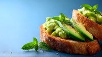 Fresh avocado toast with sliced green avocado, cucumber and mint leaves on toasted sourdough bread against blue background, healthy breakfast concept.