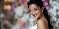 Young Asian bride with elegant updo hairstyle and white wedding dress smiling warmly at camera with soft bokeh floral background for bridal portraits.