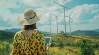 Young woman in yellow floral dress and sun hat looking at wind turbines on green hillside while holding mobile device, representing sustainable energy and modern lifestyle.