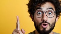 Young bearded man with glasses showing surprised expression and pointing finger up against yellow background, demonstrating eureka moment or great idea.