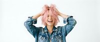 Young woman with pink shoulder-length hair wearing denim jacket, hands raised to head, smiling at camera against white background. Suitable for lifestyle, fashion, and youth culture content.
