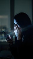 Woman with dark hair and East Asian appearance holding smartphone near face with hand, illuminated by device screen glow in dark interior setting, expressing shock or concern while using mobile technology.