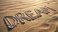 Word DREAM written with colorful shells and pebbles on golden sandy beach in warm sunlight, inspiring summer vacation concept.