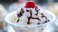 Creamy vanilla ice cream sundae topped with sweet cherry syrup and maraschino cherry in clear plastic cup, drizzled with red fruit sauce against blurred background.