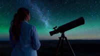 Young woman observing night sky through telescope under starry cosmos with visible Milky Way and green aurora borealis glowing on dark horizon.