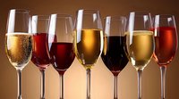 Row of elegant wine glasses filled with different varieties showing rich colors from golden champagne to deep red wine against warm background lighting.