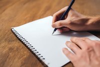 Male hand writing with black pen on blank white spiral notebook page placed on wooden desk surface for notes and planning.