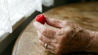 Elderly person's weathered hand holding a fresh red raspberry on wooden surface with white fabric nearby.