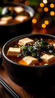 Spicy Asian soup with soft tofu cubes, seaweed and green onions in black ceramic bowl against warm bokeh lights, close-up view on wooden surface.