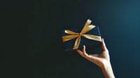 Hand holding elegant black gift box with golden ribbon bow against dark blue gradient background for special occasions and celebrations.
