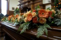 Orange gerbera daisies and roses with green ferns arranged on wooden casket at funeral service, creating somber yet beautiful floral tribute in church setting.