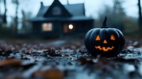 Glowing carved pumpkin with classic jack-o'-lantern face sits among fallen autumn leaves at dusk, with spooky illuminated haunted house silhouette in misty background.