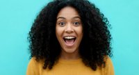 Excited young African American woman with curly hair expressing joy and surprise in yellow sweater against turquoise background for lifestyle content.