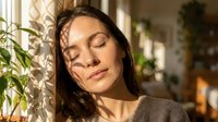 Young woman with long dark hair and olive complexion enjoying peaceful moment by window with natural sunlight, surrounded by indoor plants, expressing relaxation and wellness for lifestyle and mental health content.