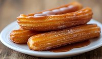 Fresh churros drizzled with warm caramel sauce on white plate against rustic wooden background, close up view showing golden crispy texture and sweet coating.