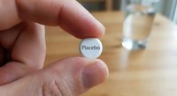 Hand holding white placebo pill tablet between fingers with glass of water on wooden table background for medical research study.