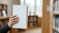 Person holding blank white paper sheet in library setting with bookshelves in background, suitable for mockups, presentations, and educational materials.