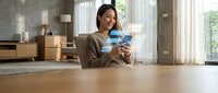 Young Asian woman relaxing at home using smartphone while drinking coffee in modern living room with natural light streaming through windows.