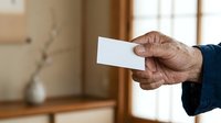 Senior man with dark skin tone holding blank white business card in hand, wearing navy blue sleeve, indoor home setting with wooden furniture and blurred background.