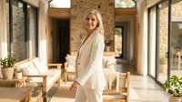 Mature woman with blonde hair wearing cream-colored blazer standing in modern brick hallway with natural light, wooden furniture and potted plants, suitable for business, lifestyle and corporate use.
