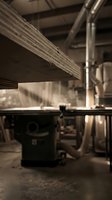 Wooden plank being cut on a table saw in a dusty carpentry workshop with warm light and industrial ventilation pipes.