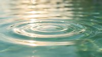 Water ripples creating concentric circles on calm turquoise surface with golden sunlight reflections. Peaceful aquatic background for wellness and meditation themes.