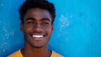 Young African American man with bright genuine smile wearing yellow t-shirt against vibrant blue wall background, expressing joy and confidence in casual portrait setting.