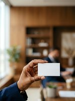 Close-up of male hand holding blank business card in modern office environment with wooden shelving and colleagues working in background.