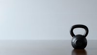 Black kettlebell weight on wooden floor against white wall background for fitness training and strength workout exercises in minimalist gym setting.