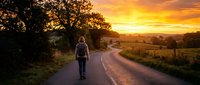 Hiker with backpack walking on rural road at golden hour sunset with warm sky and countryside landscape.