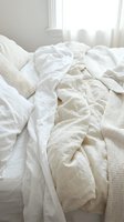 Unmade bed with white and cream rumpled linens, pillows, and knit blankets near bright window with sheer curtains, conveying comfort and relaxation for bedroom design.