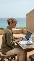Middle-aged woman with gray hair working on laptop at wooden table on coastal terrace overlooking Mediterranean sea, remote work and freelance lifestyle.