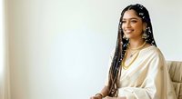 Young woman with South Asian ethnicity, long braided hair with decorative beads, wearing cream-colored satin shirt, smiling at camera against neutral background. Suitable for lifestyle, beauty, fashion, and professional content.