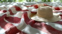 Straw sun hat on red and white striped picnic blanket with soft natural lighting and bokeh effect in background, creating summer outdoor leisure atmosphere.