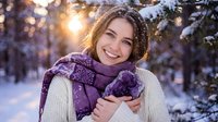 Young Caucasian woman in knitted sweater and purple scarf smiling warmly in snowy winter forest with golden sunlight filtering through snow-covered trees.