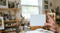 Caucasian male hand holding blank white card in creative studio workspace with shelves, plants, and natural light from window.