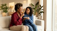 Two happy African American women sitting on beige sofa sharing smartphone and laughing together in bright modern living room with plants.