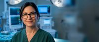 Female surgeon with glasses smiling confidently in modern operating room with surgical lights and medical equipment in background.