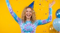 Happy young Caucasian woman with curly blonde hair celebrating in tie-dye shirt and party hat with arms raised, confetti falling on bright yellow background.