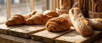 Fresh baked artisan bread loaves on rustic wooden cutting board in warm bakery kitchen with golden sunlight streaming through windows creating cozy atmosphere.